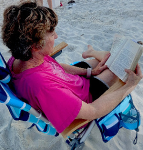 Woman in a bright pink shirt and black shorts sitting in a blue-striped beach chair on sandy beach, reading a suspense thriller.