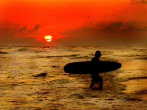 Silhouette of a man carrying a surfboard onto the beach at sunset, with a vivid orange and red sky