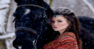Artwork from the cover of Christian romantic suspense book “Angel in Shining Armor” featuring a dark-haired woman standing beside a black horse in a snowy forest, wearing a colorful patterned garment and jeweled headpiece as snow falls around them, symbolizing faith, mystery, and quiet strength